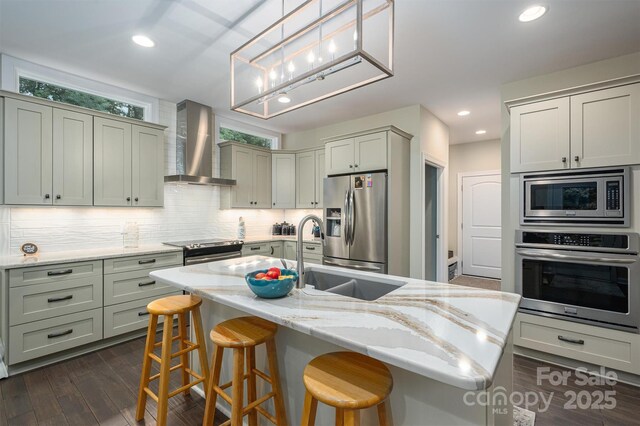 kitchen with wall chimney exhaust hood, light stone counters, decorative backsplash, a center island with sink, and appliances with stainless steel finishes