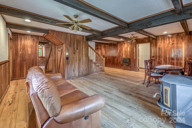 living room with a wood stove, wooden walls, ceiling fan, beamed ceiling, and light hardwood / wood-style floors