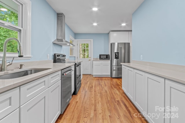 kitchen featuring light wood-type flooring, wall chimney range hood, white cabinets, appliances with stainless steel finishes, and sink