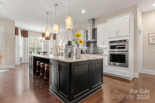 kitchen featuring a center island with sink, white cabinetry, wall chimney range hood, and double oven