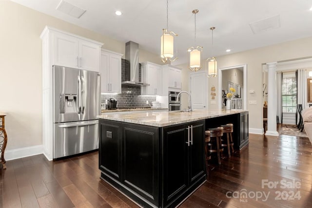 kitchen featuring pendant lighting, appliances with stainless steel finishes, white cabinetry, an island with sink, and wall chimney exhaust hood