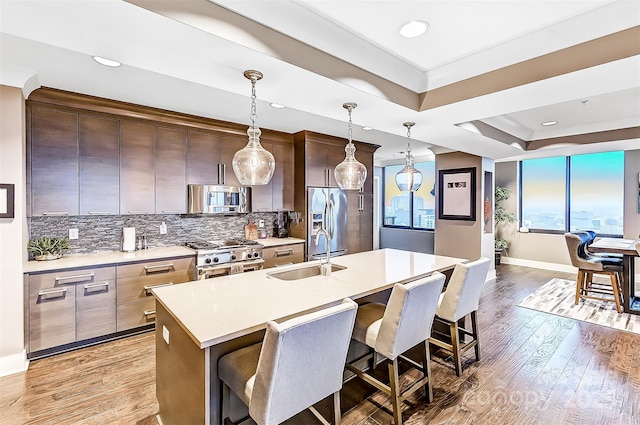 kitchen with a sink, light countertops, appliances with stainless steel finishes, an island with sink, and a raised ceiling