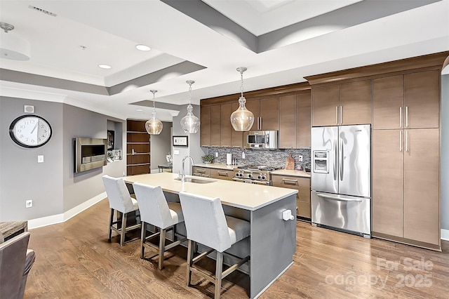 kitchen with a tray ceiling, a center island with sink, stainless steel appliances, light countertops, and a sink