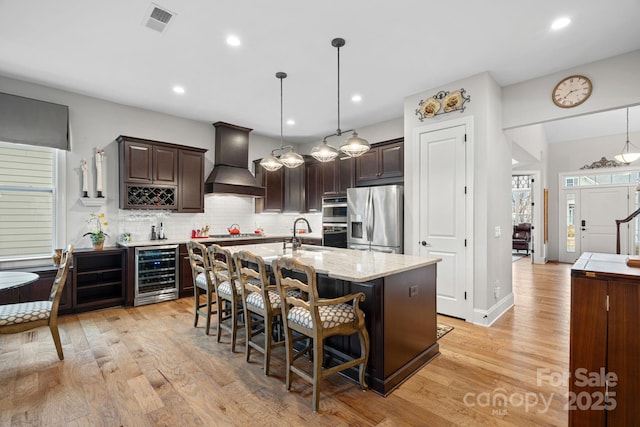 kitchen with pendant lighting, custom exhaust hood, stainless steel appliances, an island with sink, and beverage cooler