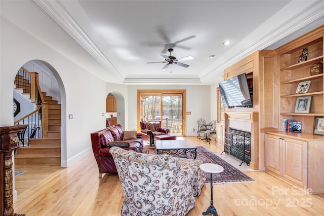 living room with arched walkways, a raised ceiling, a tiled fireplace, ornamental molding, and light wood-type flooring