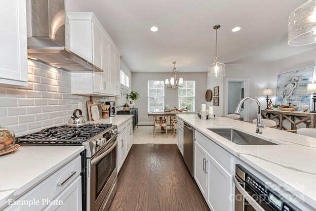 kitchen with wall chimney range hood, stainless steel appliances, hanging light fixtures, decorative backsplash, and sink