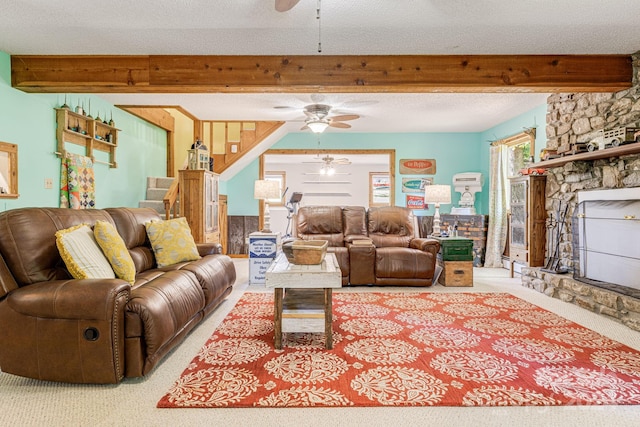 carpeted living room with a textured ceiling, ceiling fan, a stone fireplace, and beam ceiling