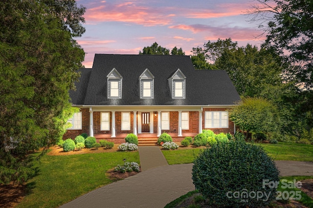 cape cod home featuring roof with shingles, a front yard, a porch, and brick siding