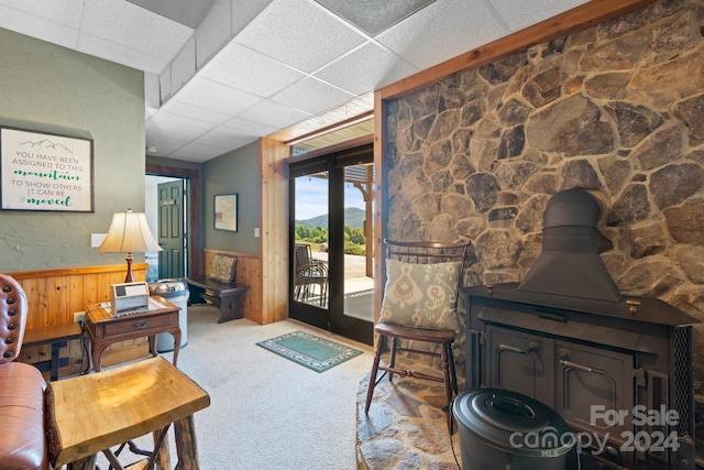 carpeted living room with a paneled ceiling, wooden walls, and a wood stove