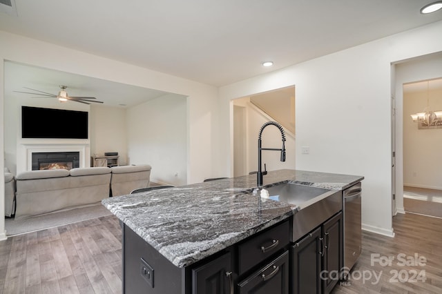 kitchen with a kitchen island with sink, ceiling fan with notable chandelier, dark stone countertops, and hardwood / wood-style floors