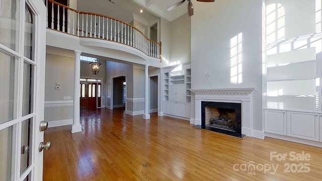 unfurnished living room with built in features, a fireplace with raised hearth, wood finished floors, ornate columns, and ceiling fan with notable chandelier