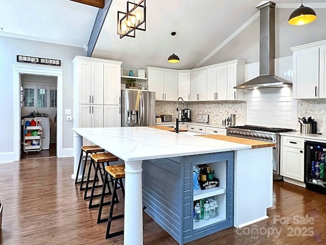 kitchen featuring appliances with stainless steel finishes, wall chimney exhaust hood, white cabinets, decorative light fixtures, and a kitchen island with sink