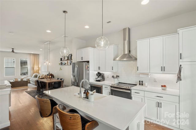 kitchen with wall chimney exhaust hood, tasteful backsplash, a kitchen island with sink, appliances with stainless steel finishes, and light hardwood / wood-style floors