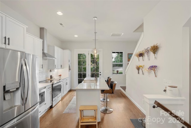 kitchen featuring stainless steel appliances, an island with sink, wall chimney exhaust hood, and light hardwood / wood-style flooring