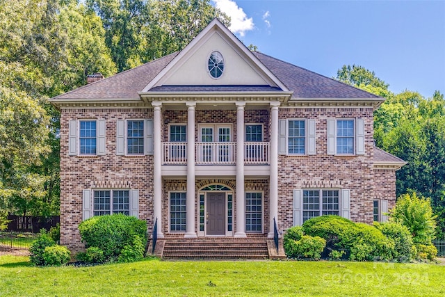 greek revival inspired property featuring brick siding, a front yard, fence, and a balcony