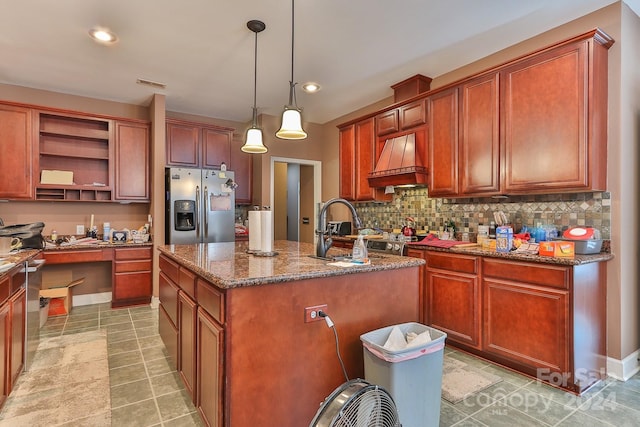 kitchen with a kitchen island with sink, custom exhaust hood, stainless steel fridge, light tile patterned floors, and decorative backsplash