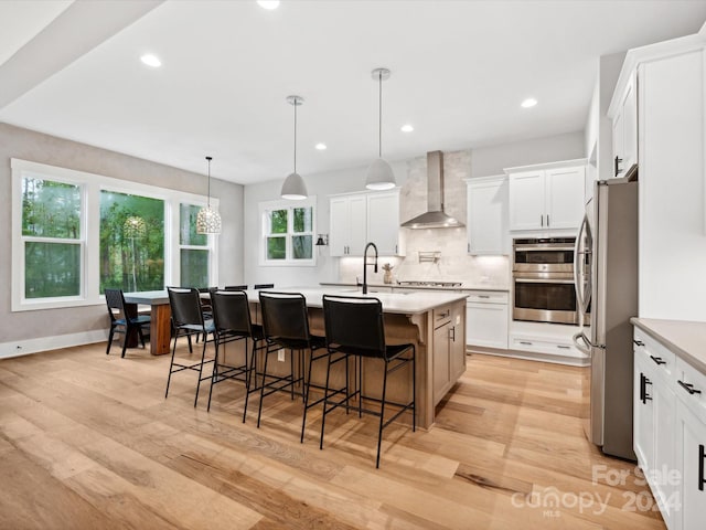 kitchen with wall chimney exhaust hood, white cabinetry, decorative light fixtures, appliances with stainless steel finishes, and a kitchen island with sink