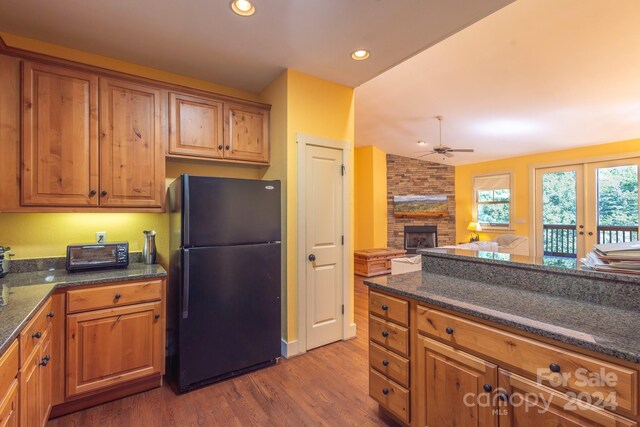 kitchen featuring hardwood / wood-style floors, a fireplace, dark stone counters, ceiling fan, and black fridge