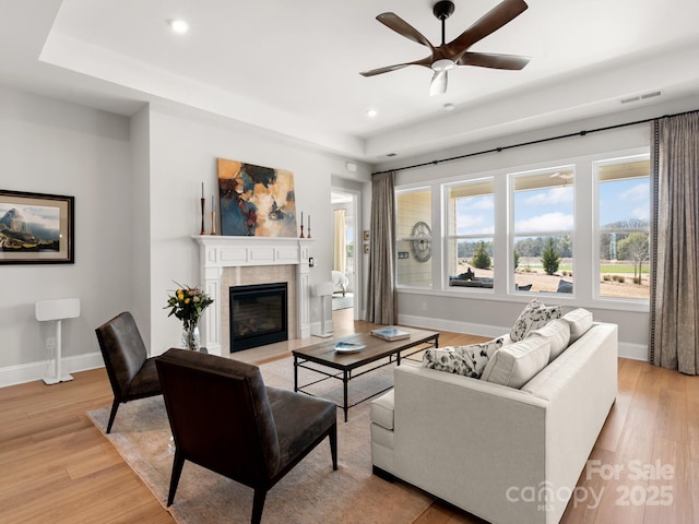 living room featuring ceiling fan, a tiled fireplace, a tray ceiling, and light hardwood / wood-style floors