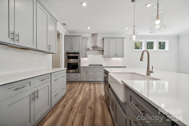 kitchen with crown molding, stainless steel appliances, gray cabinets, hardwood / wood-style floors, and wall chimney exhaust hood