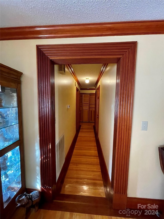 corridor featuring a textured ceiling, wood finished floors, visible vents, baseboards, and ornamental molding