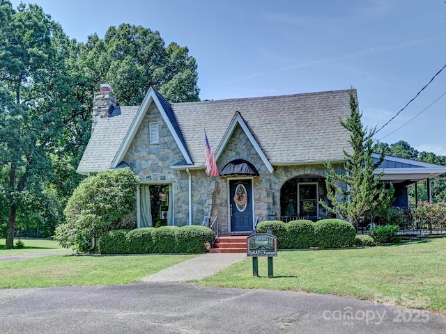 tudor home with roof with shingles, a chimney, a front yard, and stone siding
