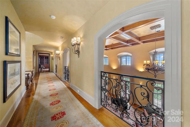 corridor featuring arched walkways, coffered ceiling, wood finished floors, a notable chandelier, and beam ceiling