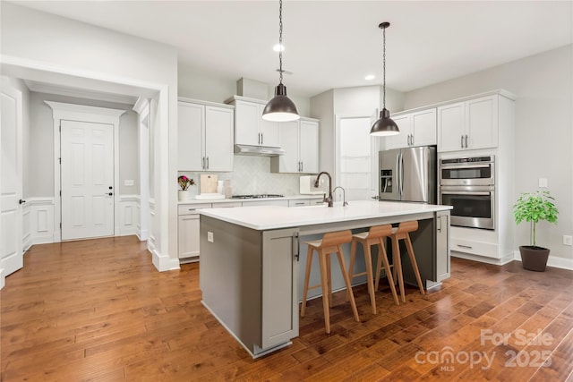 kitchen featuring a kitchen island with sink, under cabinet range hood, white cabinets, light countertops, and appliances with stainless steel finishes