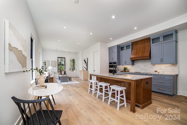 kitchen featuring light hardwood / wood-style flooring, premium range hood, backsplash, light stone countertops, and a center island with sink