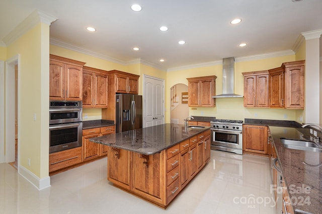 kitchen with sink, wall chimney range hood, an island with sink, appliances with stainless steel finishes, and ornamental molding