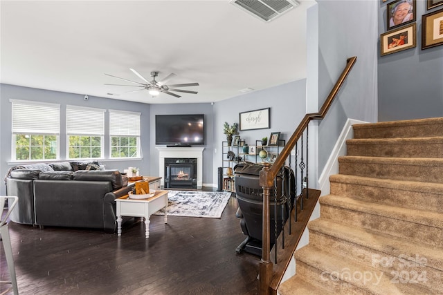 living area with dark wood-style floors, visible vents, a ceiling fan, a glass covered fireplace, and stairs