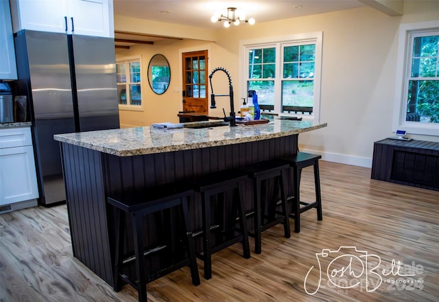 kitchen featuring stainless steel refrigerator, sink, white cabinets, light stone counters, and a center island with sink