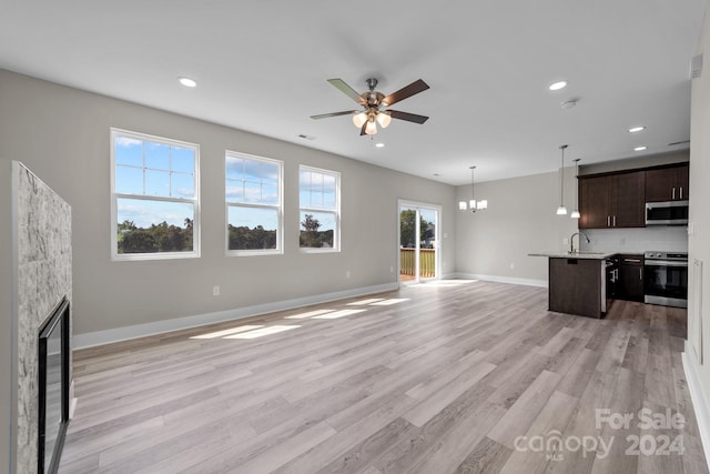 unfurnished living room with sink, ceiling fan with notable chandelier, a stone fireplace, and light wood-type flooring