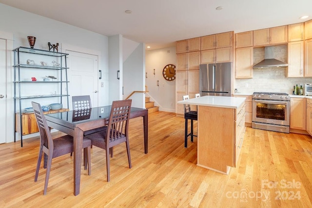 kitchen with light wood-type flooring, a kitchen island, wall chimney range hood, a kitchen breakfast bar, and appliances with stainless steel finishes