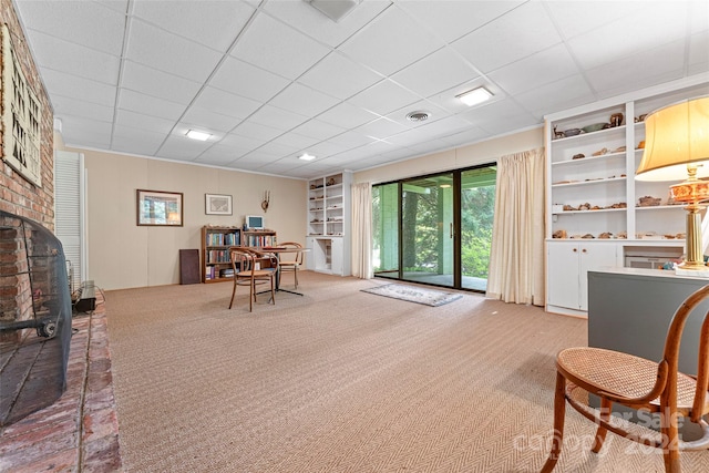 living area featuring built in shelves, brick wall, light colored carpet, a drop ceiling, and a brick fireplace