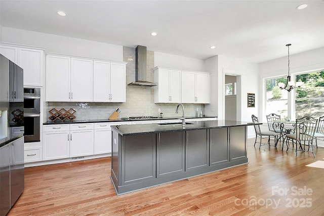 kitchen featuring pendant lighting, wall chimney range hood, sink, white cabinetry, and a center island with sink