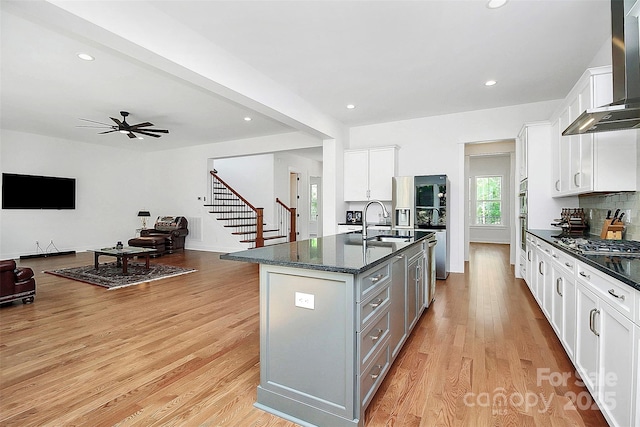 kitchen featuring wall chimney range hood, light hardwood / wood-style flooring, gray cabinets, an island with sink, and white cabinets