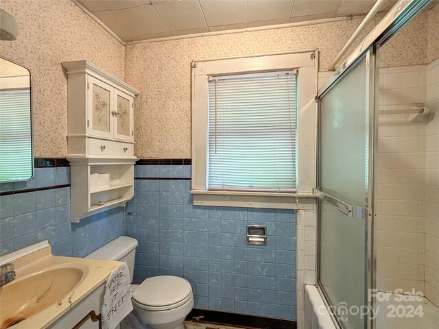 full bathroom with combined bath / shower with glass door, toilet, vanity, and tile walls