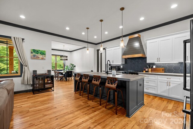 kitchen featuring pendant lighting, premium range hood, an island with sink, and white cabinets