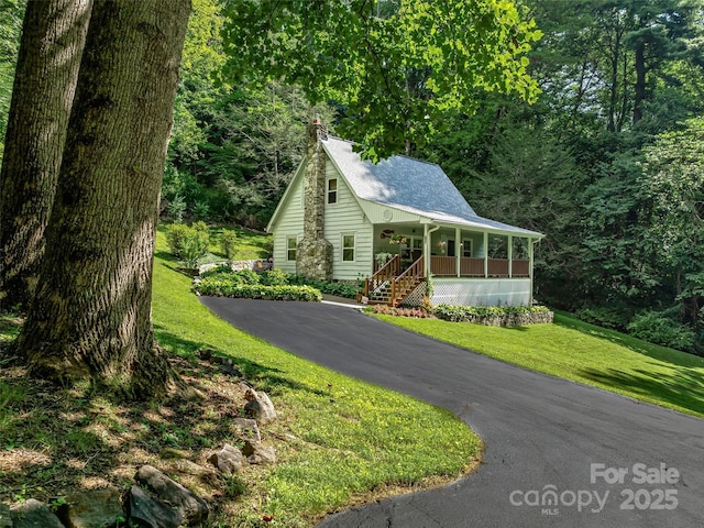 view of front of property featuring aphalt driveway, roof with shingles, a porch, a wooded view, and a front yard