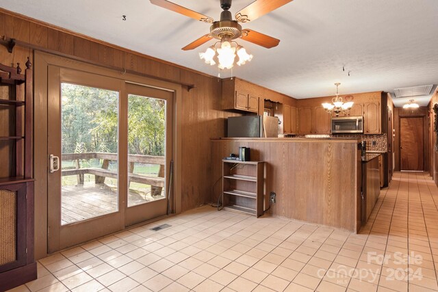 kitchen with light tile patterned floors, stainless steel appliances, wood walls, and ceiling fan with notable chandelier