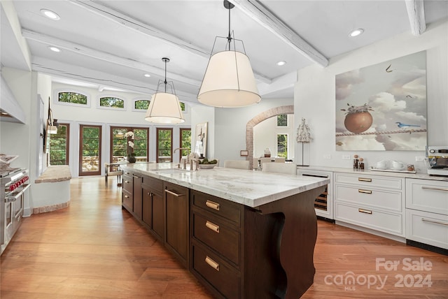 kitchen featuring light wood-type flooring, light stone counters, sink, an island with sink, and beam ceiling