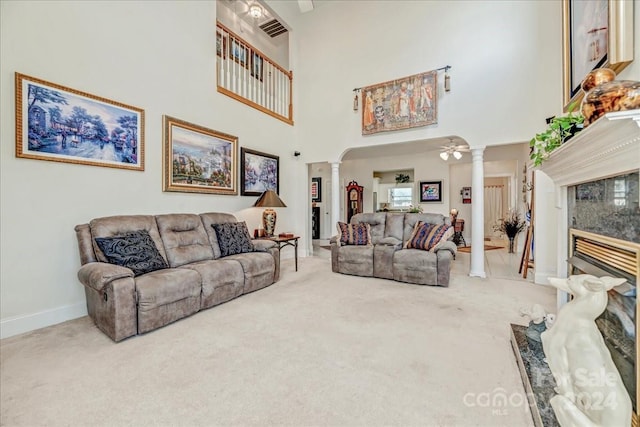 carpeted living room featuring a high end fireplace, a towering ceiling, ornate columns, and ceiling fan