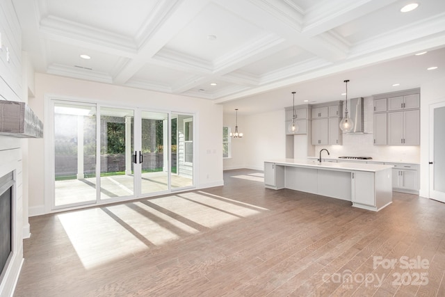 kitchen featuring light countertops, wall chimney exhaust hood, beamed ceiling, gray cabinets, and an island with sink