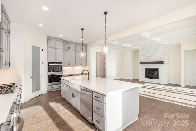 kitchen with sink, hardwood / wood-style flooring, appliances with stainless steel finishes, hanging light fixtures, and a center island with sink