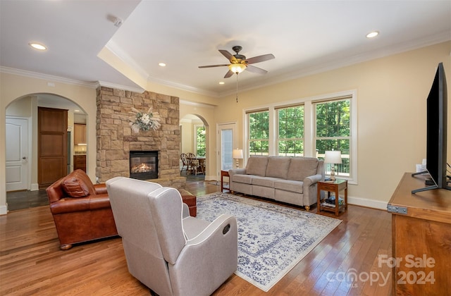 living area featuring arched walkways, light wood finished floors, ornamental molding, a stone fireplace, and baseboards