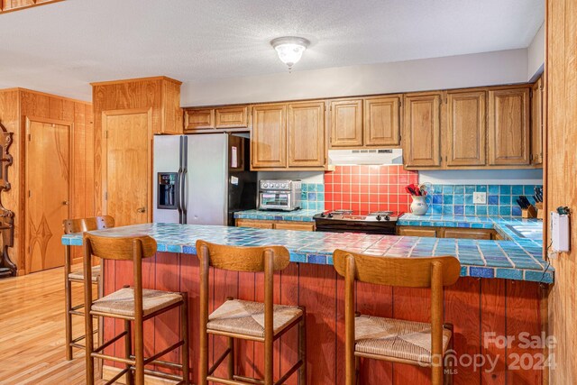 kitchen with light wood-type flooring, tile countertops, stainless steel fridge, a breakfast bar area, and tasteful backsplash