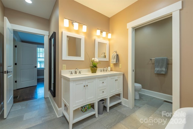 full bath with baseboards, toilet, double vanity, stone tile floors, and a sink