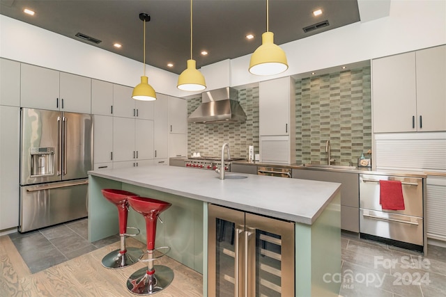 kitchen featuring built in refrigerator, wall chimney exhaust hood, wine cooler, decorative backsplash, and a center island with sink