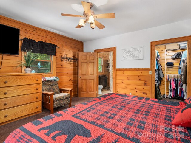 bedroom with wooden walls, carpet flooring, ceiling fan, a closet, and ensuite bathroom